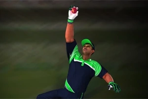 Un jucător de cricket sărind pentru a prinde o minge, purtând un echipament albastre cu verde și o șapcă asortată